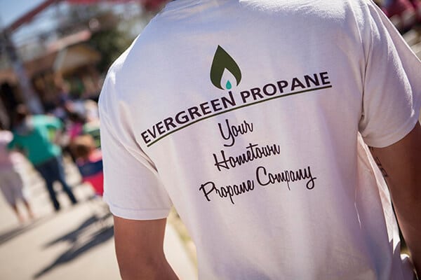 Person wearing a white shirt with Evergreen Propane - Your Hometown Propane Company printed on the back. Outdoors, with blurred people and pavement in the background.