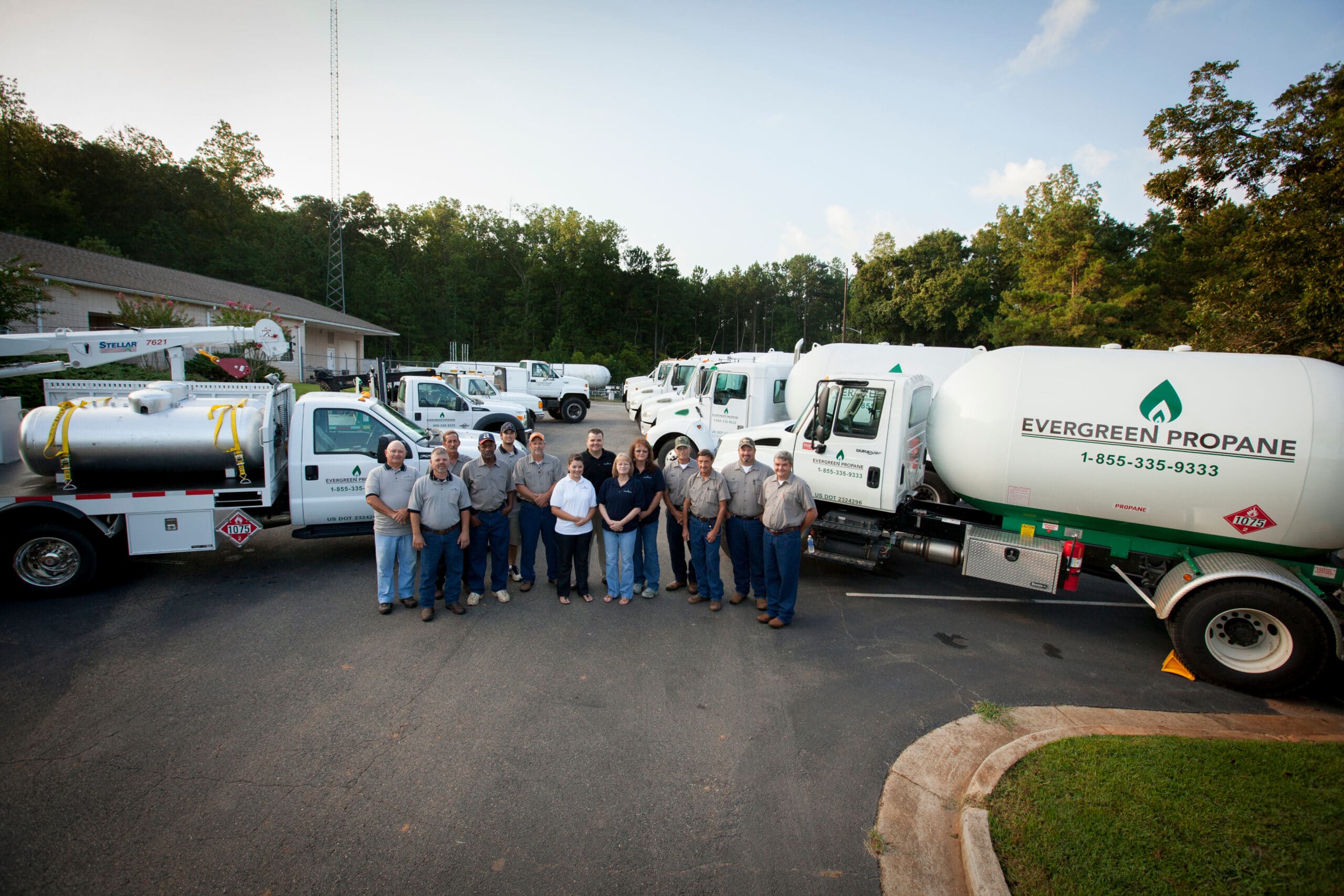 A group of people stands in front of several Evergreen Propane trucks in a parking lot, surrounded by trees and buildings, on a sunny day.