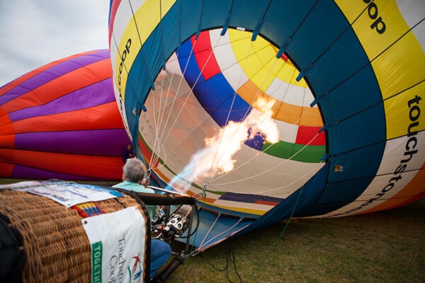 A view from behind a wicker hot air balloon basket as the burner fires into the colorful fabric envelope, which is being inflated on a grassy field. Another large purple and orange balloon is visible in the background under a cloudy sky.