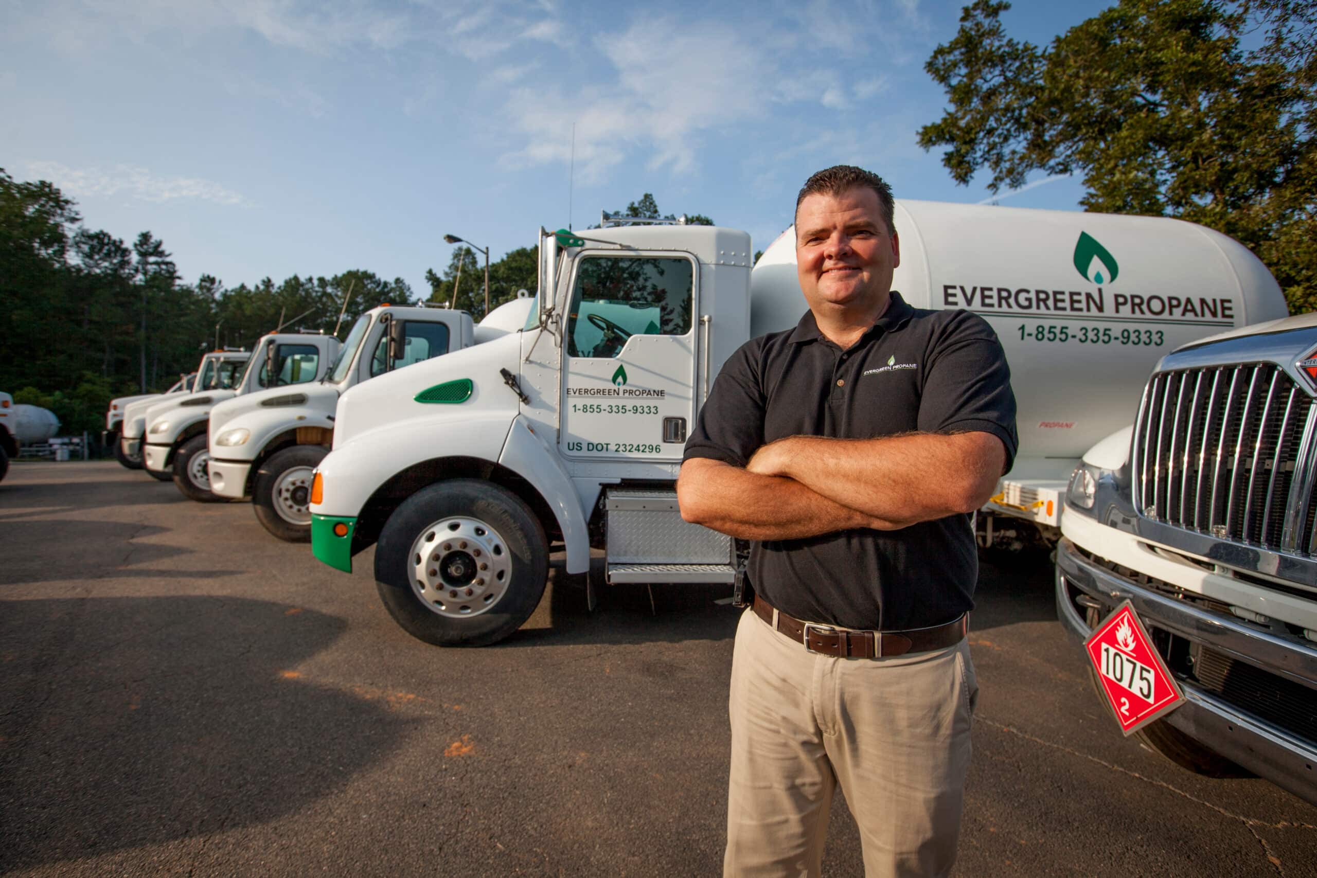 A man wearing a black shirt stands with his arms crossed in front of multiple white Evergreen Propane trucks lined up on a sunny day.