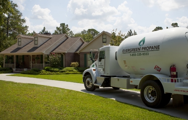 A white Evergreen Propane delivery truck is parked on the driveway in front of a brick house with a porch, surrounded by grass, trees, and a partly cloudy sky.