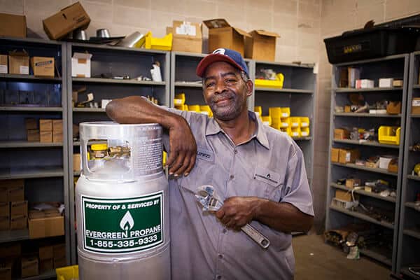 A man in a work shirt and cap stands in a supply room, smiling and holding a wrench, with one arm resting on a propane tank labeled Evergreen Propane. Shelves with tools and parts are in the background.